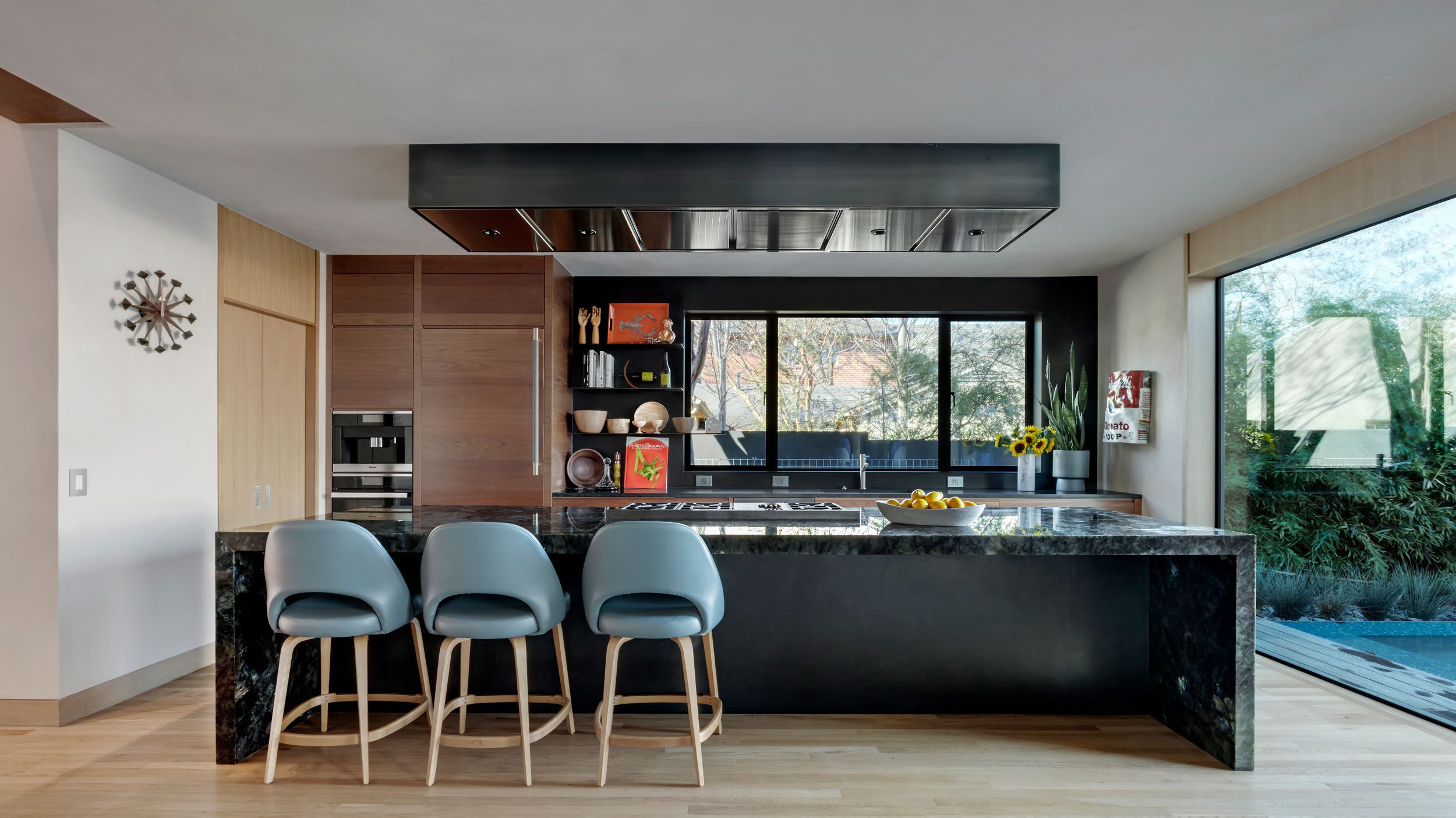 Milton-0378-hero A modern kitchen with a black marble island, three blue barstools, built-in wooden cabinets, and large windows overlooking greenery.
