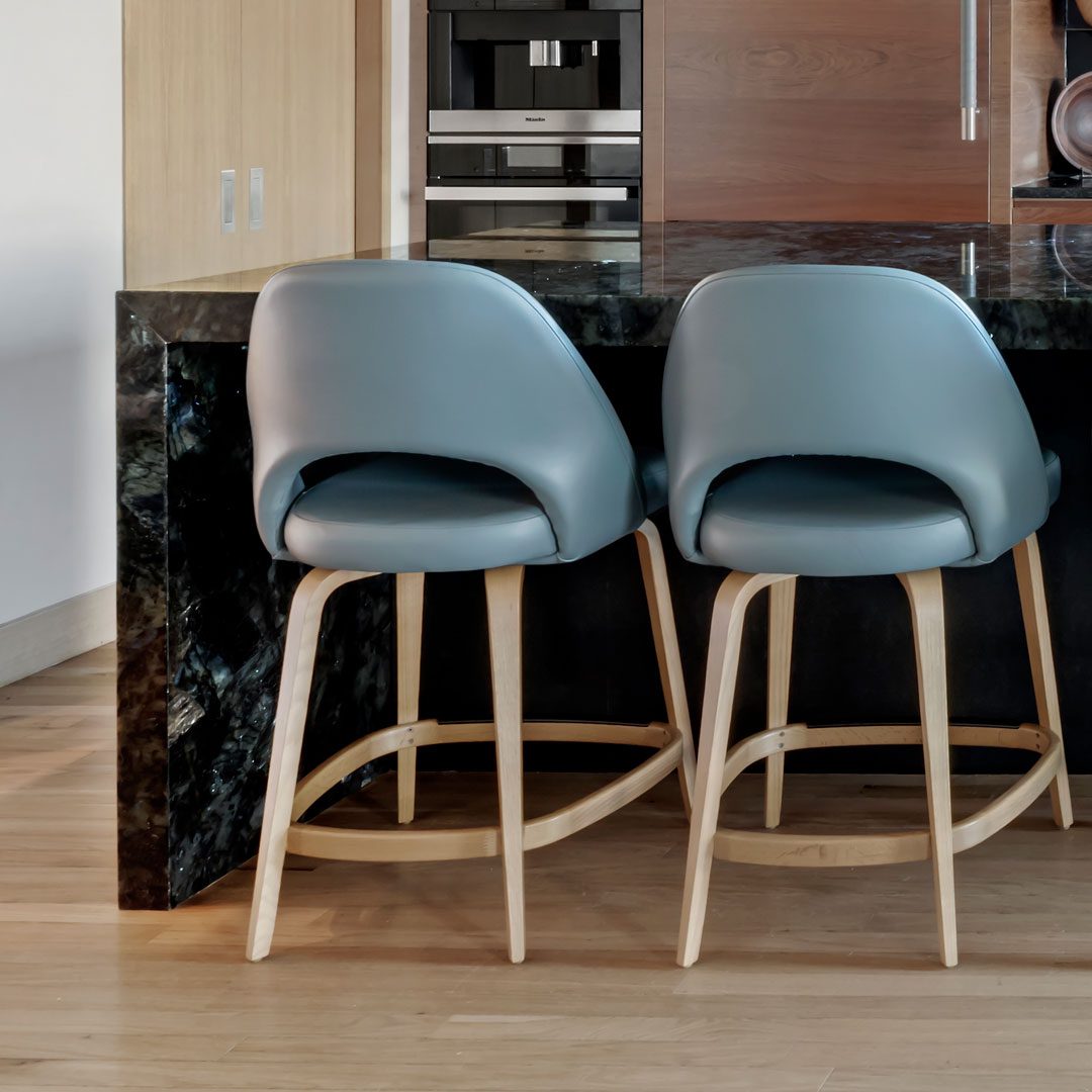 milton-1 Two modern blue barstools with wooden legs sit at a dark marble kitchen counter with built-in cabinets and appliances in the background.