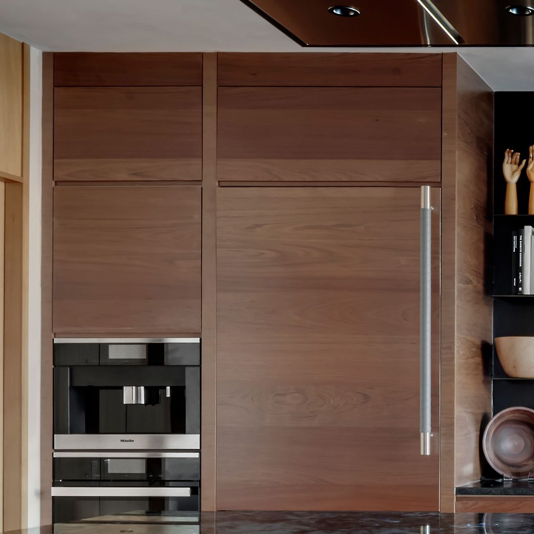 milton-2 Built-in stainless steel coffee machine and refrigerator with wood paneling, adjacent to shelves holding decorative items and bowls.