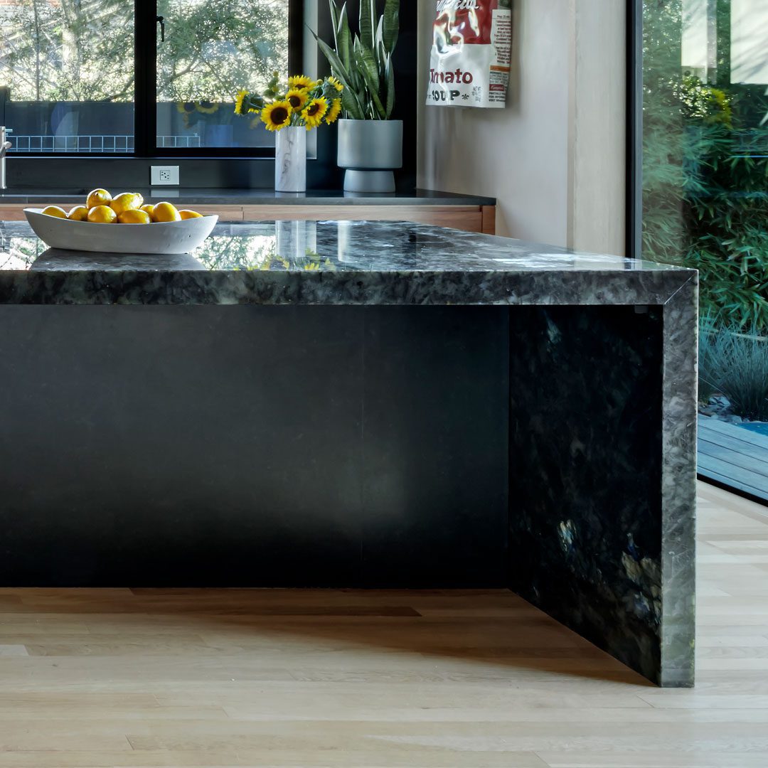 milton-mbl-2 A modern kitchen with a dark marble island countertop, a bowl of lemons, and a view of greenery through a glass door.